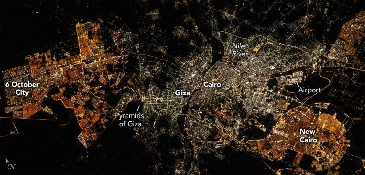 The dark image is filled with city lights. 6 October city shows up in amber lights on the left. A dark patch between that and Giza is labeled pyramids of Giza. A dark line, the Nile River separates the area of white light, Giza Left and Cairo right, in the center of the image. In the bottom right is more amber lighting and it it labeled New Cairo. The airport is just above New Cairo.