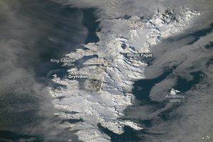 A snowy mountain range on the sea, the right side of the image is deeply shadowed. Mount Paget is in the center of the range and seems to be the tallest peak. Grytviken and King Edward point share a region on the coastline. Annenkov Island is situated in the shadowy ocean on the right side of the image.