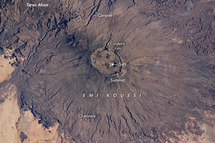 The deep brown color of the Emi Koussi stand out from the beige landscape it stands out from. Rising from the beige sands in the lower left and lower right the land is cragged with channels and even a canyon at the top center of the image. At the Summit the crags give way to a relatively smooth surface inside the Caldera rim. Near the center of the caldera is a visible, albeit fairly small, white spot labeled salt. There is a label, Tarso Ahon at the very top left of the image.