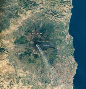 Mount Etna centers in the image with the blue sea bordering the right side of the image. A plume emanates from the center of the mountain and streams to the bottom right near the town of Catania. Multiple cones lead away from the Caldera in a straight line down and to the left.