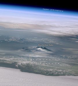 Image shows the Pacific Ocean in the foreground of the Kamchatka Peninsula. The mountain peaks of Klyuchevskaya Sopka are visible inland, cutting through some light cloud cover. In the upper left corner a stretch of reflective Earth is labeled the Sea of Okhotsk with the blue band of upper atmosphere visible across the top of the image, giving way to the dark of space in the upper right corner.