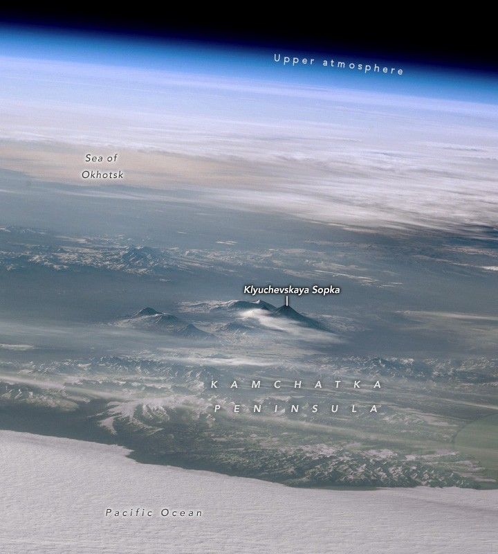 Image shows the Pacific Ocean in the foreground of the Kamchatka Peninsula. The mountain peaks of Klyuchevskaya Sopka are visible inland, cutting through some light cloud cover. In the upper left corner a stretch of reflective Earth is labeled the Sea of Okhotsk with the blue band of upper atmosphere visible across the top of the image, giving way to the dark of space in the upper right corner.