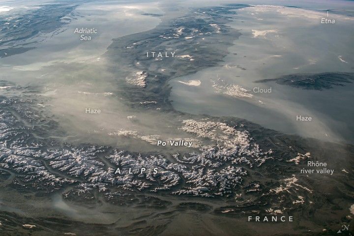 The image shows the peninsula of Italy stretching to the top of the image with the snow capped Alps in the foreground. The Adriatic Sea in the upper left gives way to haze that covers the Po Valley in a haze. Etna is labeled at the upper right.