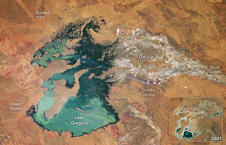 Light blue-green Lake Gregory resides amid orange brown landscape with linear dunes and Burned areas to the left and a near white shade of land webbing into the lake from the right. Labeled as A Delta of Sturt Creek. Along the lower portion of the delta is a faint smoke plume streaming left. A pop out image of the same lake in 2021 shows the lake taking up much less of the landscape.
