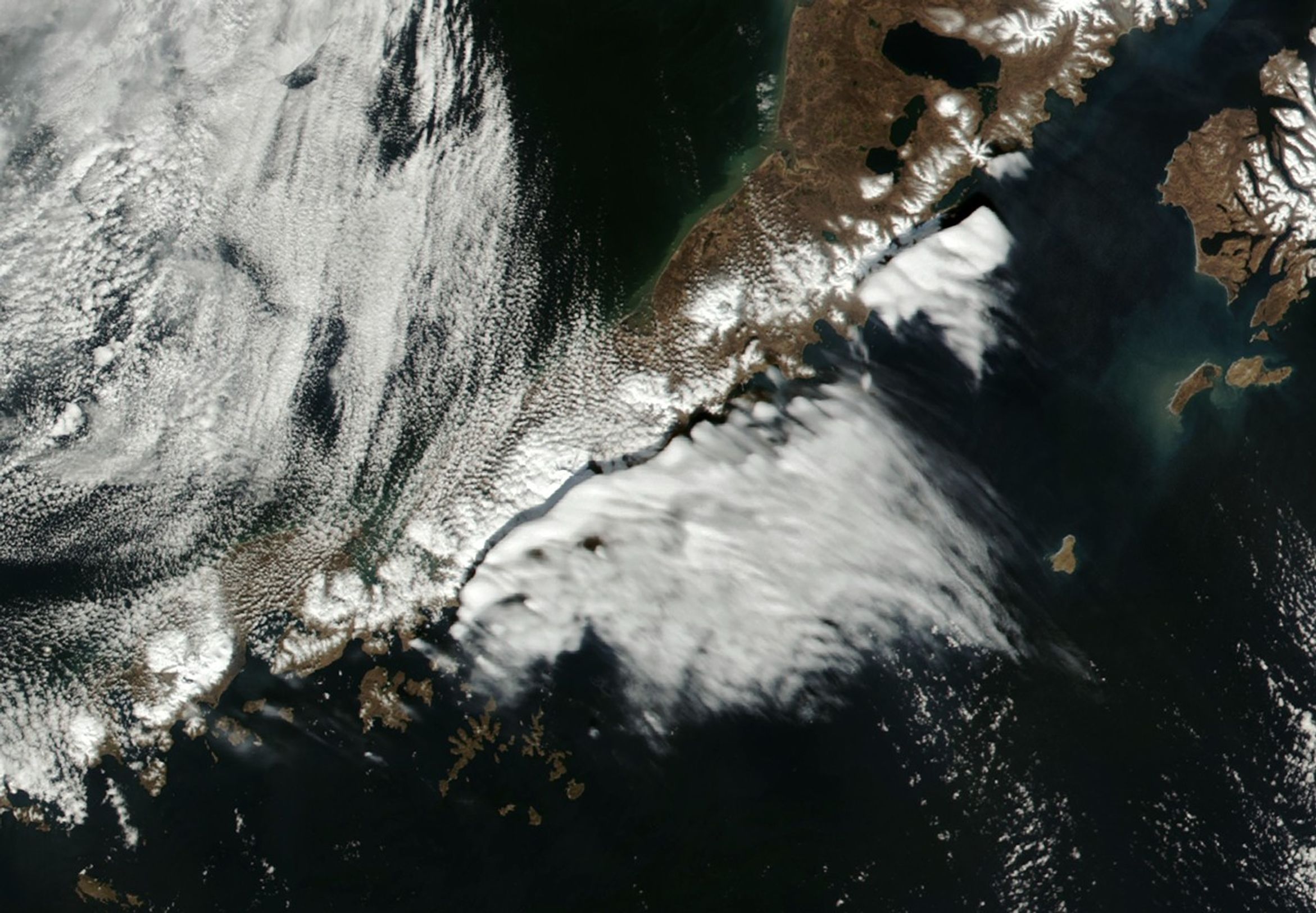 A narrow peninsula divides bodies of water, the Bearing Sea above and the Gulf of Alaska below. Two separate dense clouds occupy the air above the lower coastline, a sheet of cloud in the wake below them and thin wisps of clouds coming down from the top left corner of the image. The land is brown in color with sporadic white snow capping and the clouds bright white.