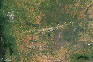 The upper left corner shows the human settlement of Calgary as a distinctly gray amorphous dot on the landscape. Along the left border of the image appears to be deep green foliage covered slightly mountainous landscape. The vast majority of the image is covered in speckled and gridded farmed fields with a distinct path of discoloration stretching horizontally across the bulk of the image labeled hail scar.