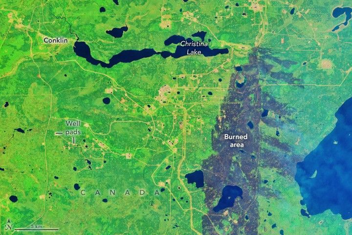 In this satellite image the majority of the landscape is vibrant green with Christina Lake stretching horizontally across the upper part of the image. At the left end is Conklin. Below that halfway down the image are well pads. To the right side of the image is a large brown region that stretches vertically into the image from the bottom. labeled burned area.