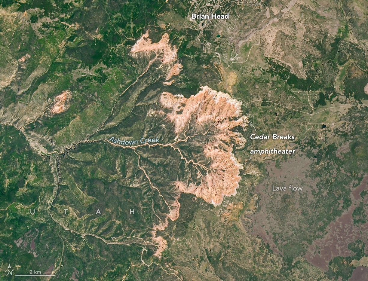 A Landsat image is centered on an orange, amphitheater-shaped escarpment cutting into darker green, forested terrain at Cedar Breaks National Monument in southwestern Utah. Light-colored and orange cliffs and ridges form a semicircular rim, with deeply eroded drainages radiating westward. A black lava flow with little vegetation is visible east of the escarpment. Trails from Brian Head, a nearby mountain and ski area, are visible to the north.