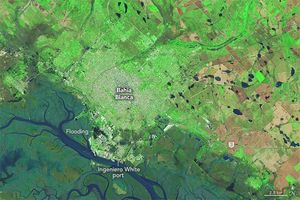 The upper portion of the image is bright green with a human settlement labeled Bahía Blanca above the river seen across the bottom of the image with wide marshes. Ingeniero White port sits along the dark blue river, and flooding is labeled between this and the city center.