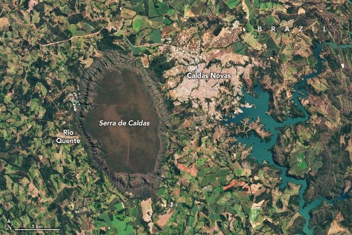 A dark brown prominent oval shaped geological feature is labeled Serra de Caldas. To the left of that is Rio Quente. To the right of the oval is Caldas Novas, a human settlement, appearing light tan, nestled between Serra de Caldas and a major branching water reservoir. The remainder of the image is varying shades of green and tan fields dotted with small spots of dense foliage.
