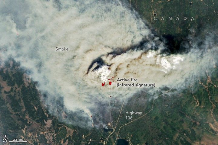 red dots, labeled active fire (infrared signature) are highlighted in the center of the densest smoke that occupies the center of the image and disperses primarily to the upper left corner of the image. Highway 106 in Canada reaches from somwhere below the smoke to the middle bottom of the image. The surrounding landscape is a mottled green