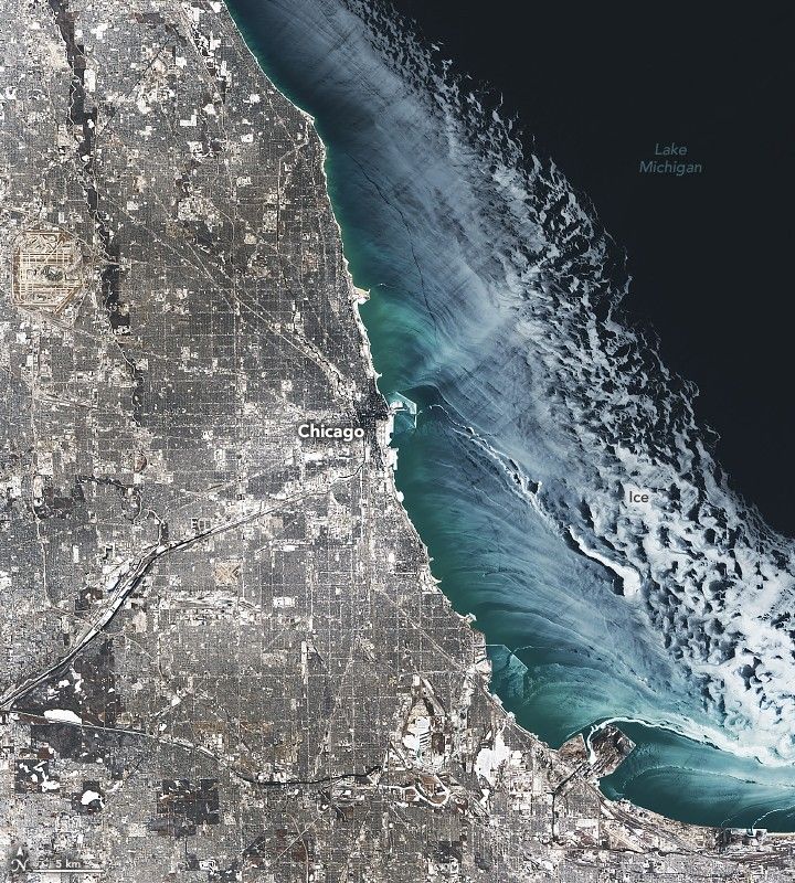 Greater Chicago is shown, left, appearing a translucent pale white color with Lake Michigan, right, also covered near the coast by a sweeping pale white ice layer. The water in the top right is ice free and appears near black, while coastal waters appear lighter and blue-green.
