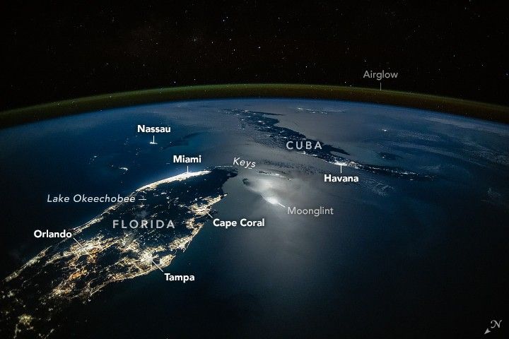 An oblique nighttime photo shows the Florida Peninsula on the left and Cuba on the middle-right, with clusters of city lights illuminating both landmasses. Moonglint shimmers across part of the ocean surface, while green airglow and the darkness of space stretch across the top of the image.