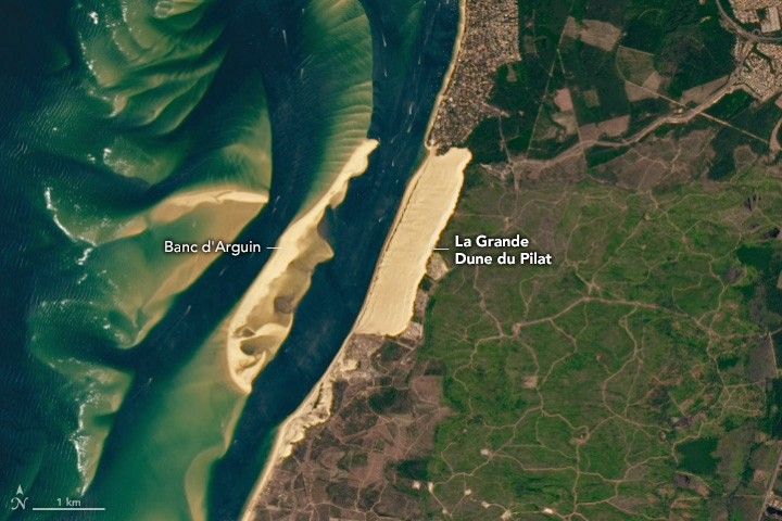 off shore is Banc d' Arguin, a sand bar that prominently sits above the water, it is surrounded by shallow covered sandbars. La Grande Dune du Pilat is a region of light beige sands on the coast opposite. The land based dune is wide and long, with green landscape to the right, human settlements above and blue waters on the left.