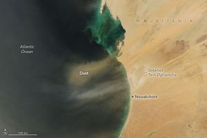 A yellow cloud of Dust is seen over the dark Atlantic Ocean off the coast of sand covered Mauritania. The capital, Nouakchott, is labeled on a the smooth curved portion of the coast. A patch of slightly different colored land, just off the coast above the capital, also seems to be putting dust into the atmosphere parallel to the yellow dust cloud, it is lighter in color than the yellow dust plume.