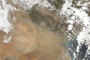 A light shade of dust in an arc sweeps across Iraq along the border with Saudi Arabia. The land below is a darker shade of beige than the dust. Iraq shows a significant brown and green shade of landscape. Saudi Arabia in the lower left appears more reddish. Kuwait City in the lower right sits on the coast, mostly obscured by cloud cover that frames the image, except for the lower left. An Najaf resides on the northern end of the dust and green region of Iraq. Al Başrah resides directly North of Kuwait City in Iraq.