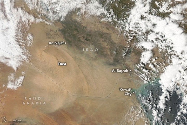 A light shade of dust in an arc sweeps across Iraq along the border with Saudi Arabia. The land below is a darker shade of beige than the dust. Iraq shows a significant brown and green shade of landscape. Saudi Arabia in the lower left appears more reddish. Kuwait City in the lower right sits on the coast, mostly obscured by cloud cover that frames the image, except for the lower left. An Najaf resides on the northern end of the dust and green region of Iraq. Al Başrah resides directly North of Kuwait City in Iraq.