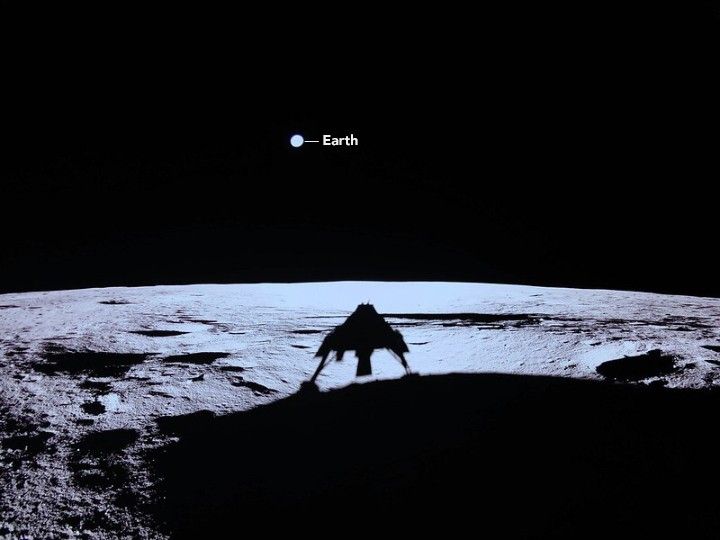 The upper portion of the image is filled with black space and a blue-gray circle labeled Earth. The foreground shows the expanse of the bright gray and dark shadowed moon, including the shadow from the lander taking the photo which has two distinct legs.
