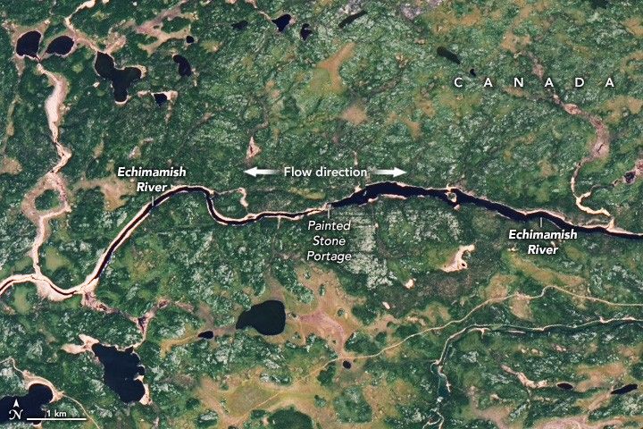 The Echimamish River flows horizontally across this image. At the image center a painted stone portage is labeled, from which, the water on the left side of the river is considerably more narrow that on the right. The surrounding landscape is a mix of green shades with sporadic lakes.