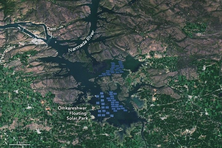 Blue rectangles sit in the midst of a dammed reservoir. The dam is labelled in the upper left and the Narmada river enters the photo from the top center. The land below the reservoir is largely green with foliage, and the upper portion of the image is dark gray and rocky.