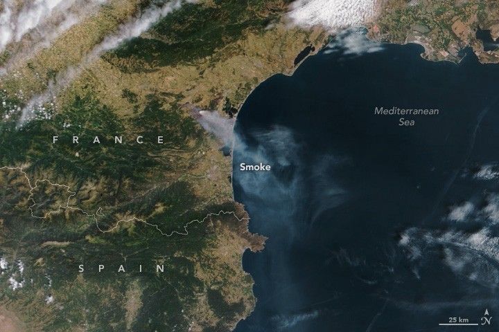 The landscape of France and Spain occupies the upper and left portion of the image, with Spain at the bottom of the image. The landscape is a mix of green foliage and yellowish brown, at the edge of where these two colors meet a plume of smoke emanates and reaches toward the rich blue of the Mediterranean Sea, occupying the lower right of the image.