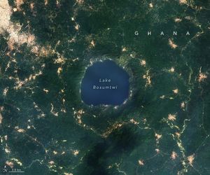 Lake Bosumtwi, a rich blue hue, is surrounded on all sides by elevated land rich green foliage. There are a patchwork of tan color human settlements within the foliage, and one major region is built up in the top left that seems to be building in the direction of the lake.