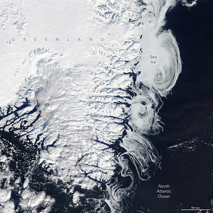 The image shows bright white snow covered Greenland, occupying the upper and left portion of the image with the near black waters of the North Atlantic Ocean in the lower and right side of the image. Along the coastline, right of Greenland, swirls of Sea ice are visible all the way to the tip.