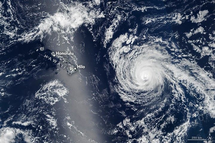 The image shows the deep blue waters of the Pacific Ocean around Hawaii. The islands have been outlined in white on the image and cities of Hilo and Honolulu labeled. A swirl of bright white clouds is off the coast to the right of the islands and sporadic cloud coverage occupies much of the image.
