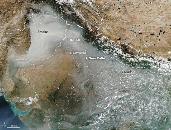 A Satellite image of India, a swath of smoke follows the contour of the snow capped Himalayas obscuring Punjab, Haryana, New Delhi and more of India before dispersing amid clouds at the bottom right of the image.