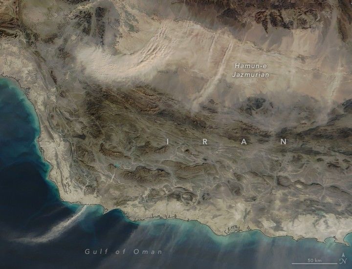 Multiple plumes of dust stretch out over the Gulf of Oman at bottom of the image from Iran and the Hamun-e Jazmurian occupying the top of the image. It is a stretch of land that seems relatively flat and without vegetation. lighter in color than the surrounding region.