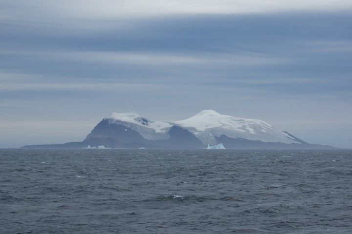 This photograph shows Candlemas Island appearing slightly hazy in the distance, with a wavy ocean in the foreground and a partly cloudy sky in the background. The island is dark and rocky at its base and topped with glacial ice.