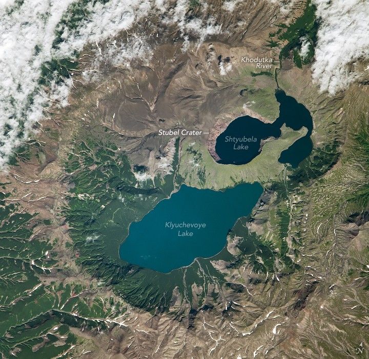 In a depression surrounded by mountains on all sides are Klyuchevoye Lake and Shtyubela Lake, the former of which is a light shade of blue and larger than the dark Shtyubela Lake. Shtyubela Lake is also connected to Khodutka River which exits the image up and to the right. The landscape varies from deep green foliage, primarily to the lower left and around the river to bare beige rock. Thin streaks of snow appear in the many valleys around the Stubel Crater that holds these lakes.