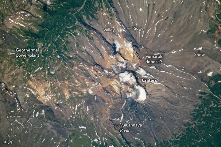 The image shows the volcano, the image is dominated by the dark tan features of a volcanic summit, at the base in the upper left and lower right, the tan gives way to deep green foliage. The vulkannaya river is highlighted at the bottom of the image. White snow is smattered across the volcano ridges and valleys. The craters at the top are snow covered and there is a golden colored soil labeled fumarole deposit? A geothermal power plant is labeled in the valley in the upper left.