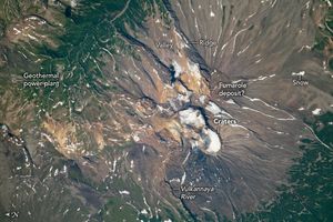 The image shows the volcano, the image is dominated by the dark tan features of a volcanic summit, at the base in the upper left and lower right, the tan gives way to deep green foliage. The vulkannaya river is highlighted at the bottom of the image. White snow is smattered across the volcano ridges and valleys. The craters at the top are snow covered and there is a golden colored soil labeled fumarole deposit? A geothermal power plant is labeled in the valley in the upper left.