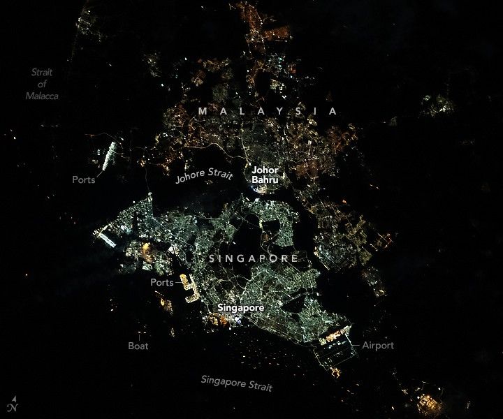 The Johore Strait separates Singapore (south) from Peninsular Malaysia (north). The urban area of Singapore encompasses the country’s main island, Singapore Island, in addition to numerous smaller islands scattered throughout the Johore and Singapore straits. Two illuminated bridges, the Johor Causeway in the central region and the Second Link toward the west, connect the two countries. The Strait of Malacca on the top-left side of the image appears dark, lacking much light from boats or land, whereas the right side is lined with small villages of mainland Malaysia.