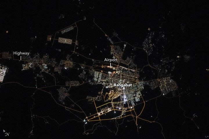 The image shows the bright lights of the city and major roadways of Ashgabat. A highway entering the city and surrounding streets are also somewhat illuminated. The Airport is centered in the image with the city mostly residing below and right of it. A significant amount of the land surrounding the city is completely dark.