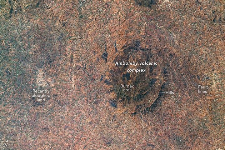 Most of the landscape in the image is light brown, indicating a lack of forest cover. In contrast, greener areas farther west (along the left third of the image) retain more of the original forest of Madagascar’s central highlands. Amid that region is a light colored patch of land labelled recently ploughed land.  Fault lines run vertically on the image to the right of the volcanic complex. Hills separate the fault from the complex and a black area in the middle of it is labeled the burned area.