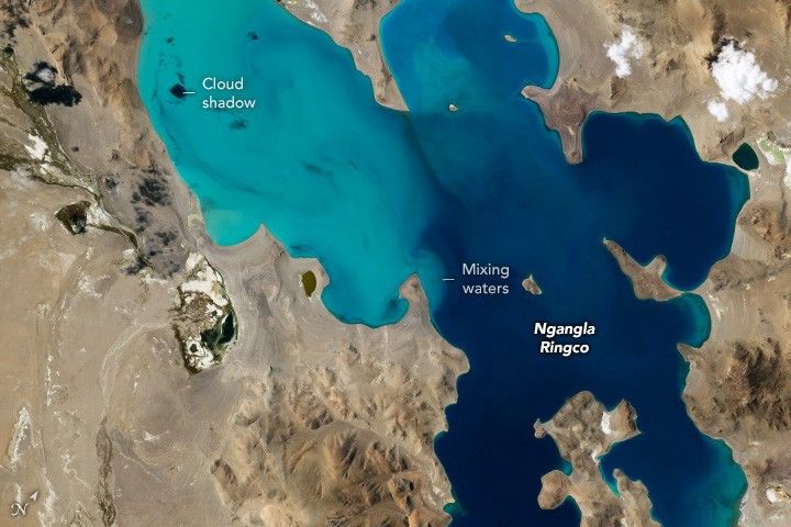 Turquoise waters are surrounded by sandly landscapes. A dark spot is shown as a cloud shadow in the upper left. The waters in the lower right are darker and distinctly swirl into a mix where they meet. Label Ngangla Ringco is over the darker blue waters.