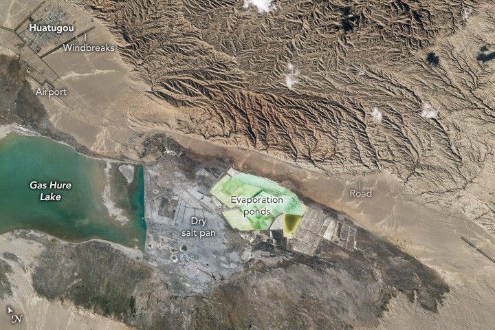 Gas Hure Lake is centered on the left of the image below an Airport and windbreaks around the town of Huatugou in the top left corner.  The top center of the image appears to be some mountainous terrain, a road separating that from the Dry salt pans and evaporation pond in the lower center portion of the image. The Evaporation ponds are a pale seafoam color. The immediate land around it is darker in color. 