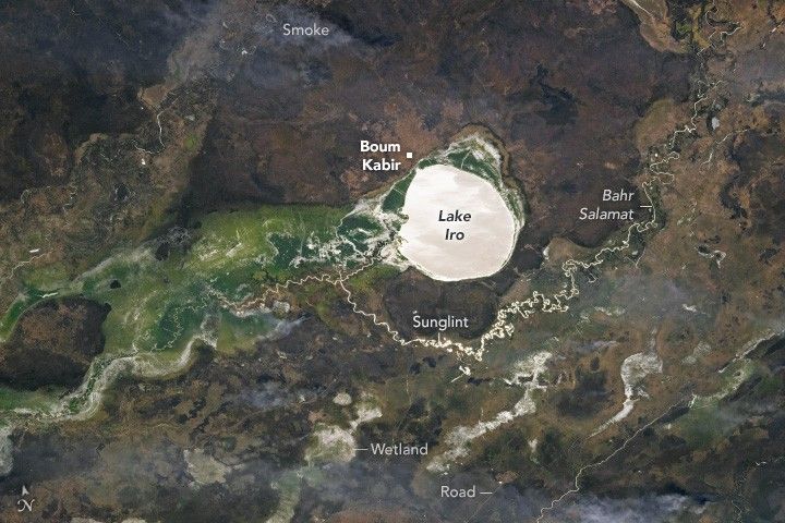 I near white Lake Iro centers the image just below Boum Kabir. Smoke is in the upper left corner, but appears only a wispy haze. The region left of the lake is a vibrant green. Wetlands and a road are labeled to the lower center of the image, with the road being visible as a thin discolored line amid the green/brown landscape. Bahr Salamat is a river meandering through the image, vibrant with sunglint below lake Iro.