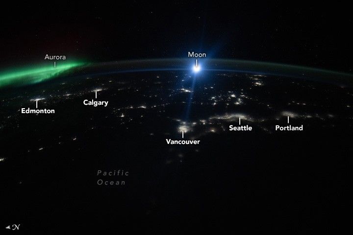 The West coast of the United States and Canada at night shows the city lighting of Portland, Seattle, Vancouver, Calgary, and Edmonton. The green lights of the aurora is visible on the left edge of the image.