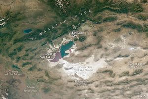 The Wasatch Range of mountains, covered in a deep green that becomes gradually more sage as it blends with the beige valleys below, span horizontally across the top of the image. Below that is Salt Lake City, nestled just above the Great Salt Lake. Below that is the Great Salt Lake Desert, which includes the Bonneville Salt Flats, all of which is white to off white. To the right is Great Basin National Park amid the gray brown dusty landscape. In the lower left are the Craters of the Moon and the Snake River Plain. The landscape in this portion of the image appears more true brown than the gray-brown of the lower right.