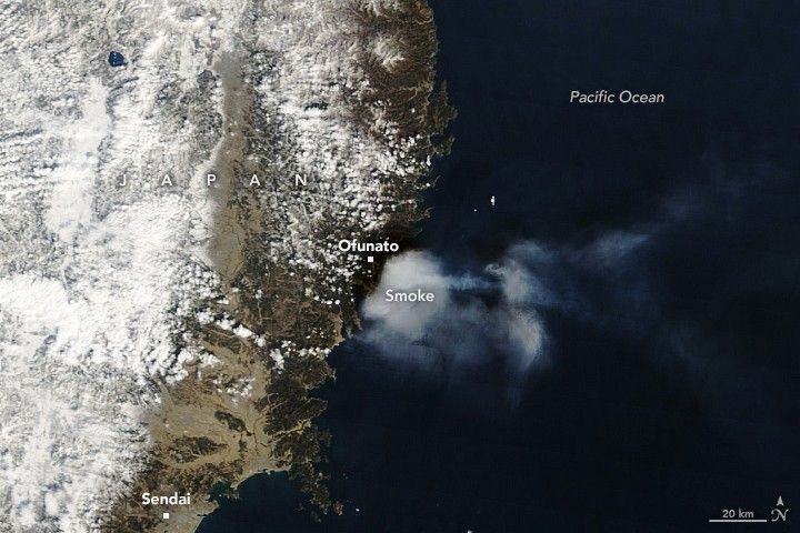 Smoke emanates from the coastline below the coastal city of Ofunato over the deep blue Pacific Ocean on the right. The land on the left is largely covered by snow further inland and reflects a vibrant white. The city of Sendai is labelled at the bottom left of the image.