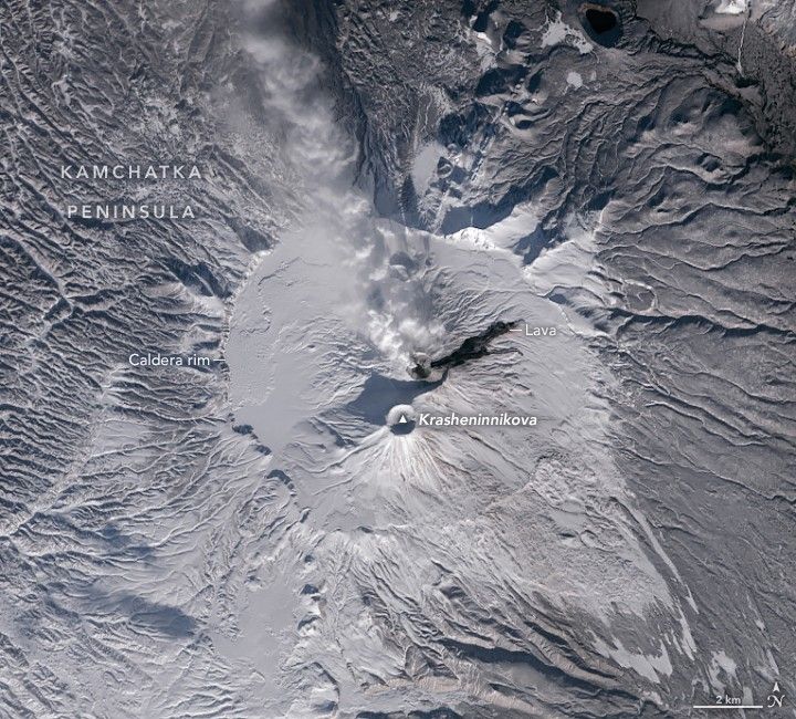 a snow covered volcano peak has an interior ring of smooth snow labeled the caldera rim, a dark streak labeled lava to the upper left and the craggy peaks and valleys of the Kamchatka peninsula surrounding.