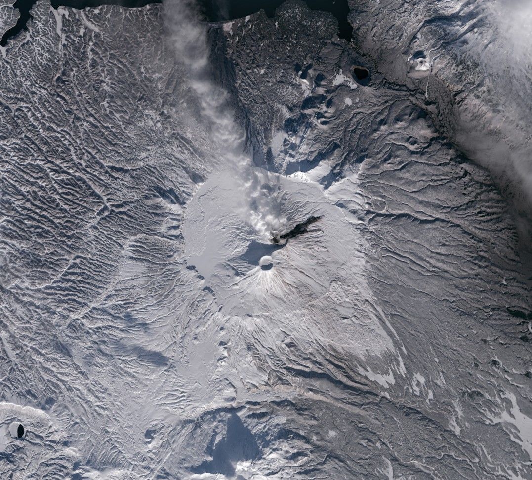 A satellite image focuses on the Krasheninnikova volcano on the Kamchatka Peninsula. The snow-covered summit caldera contains two small, round craters. A plume billows to the northwest and a dark lava flow runs to the northeast from the northern crater. The surrounding slopes appear to be covered in a thin layer of white snow.