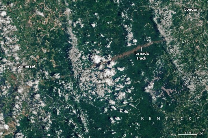 The towns of Somerset, left, and London, right, in Kentucky are divided by a large swath of deep green foliage. Between the two towns a line of brown land disrupts the foliage and is labeled as a tornado track. Sporadic cloud cover covers much of the image, and the two towns show varied forms of land use including farmed fields in tan shades.