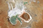 A satellite image centers on Lake Eyre in the Australian outback. Two large lobes in the southern part of the lake contain water. The one to the left appears green, and the one on the right is a rusty red. The lakebed to the north is bright and appears dry. The surrounding desert appears in shades of orange and tan.