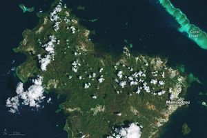 The image features an island with sporadic cloud coverage and primarily covered in a neutral green foliage. The settled parts of the island are primarily around the coast and appear white. This includes Mamoudzou on the right side of the image. The surrounding waters are a moderate navy blue except a thick line in the upper right parallel to the shore which is turquoise and labeled as coral reef.