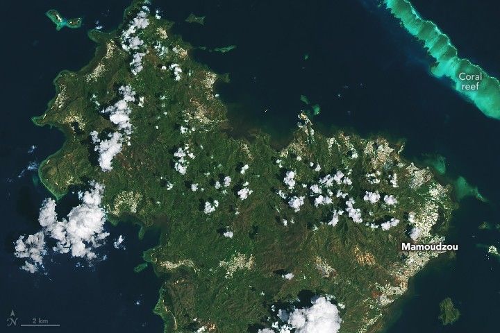 The image features an island with sporadic cloud coverage and primarily covered in a neutral green foliage. The settled parts of the island are primarily around the coast and appear white. This includes Mamoudzou on the right side of the image. The surrounding waters are a moderate navy blue except a thick line in the upper right parallel to the shore which is turquoise and labeled as coral reef.