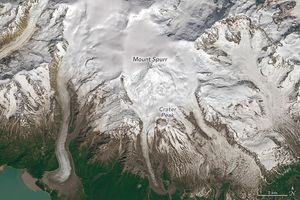 Snow covered mountainous terrain, Mount Spurr label is centarlly located in the image. Crater peak is lower and slightly right of that and is where the brown land starts to peak through the snow in streaks. In the lower left corner there appears to be a body of blue-green water.